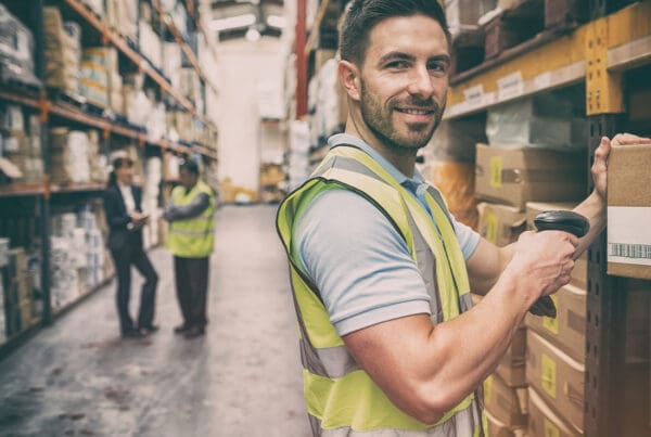 Warehousing and Logistics Insurance - Portrait of a Warehouse Worker Scanning a Box While Smiling With Fellow Colleagues Blurred in the Distance